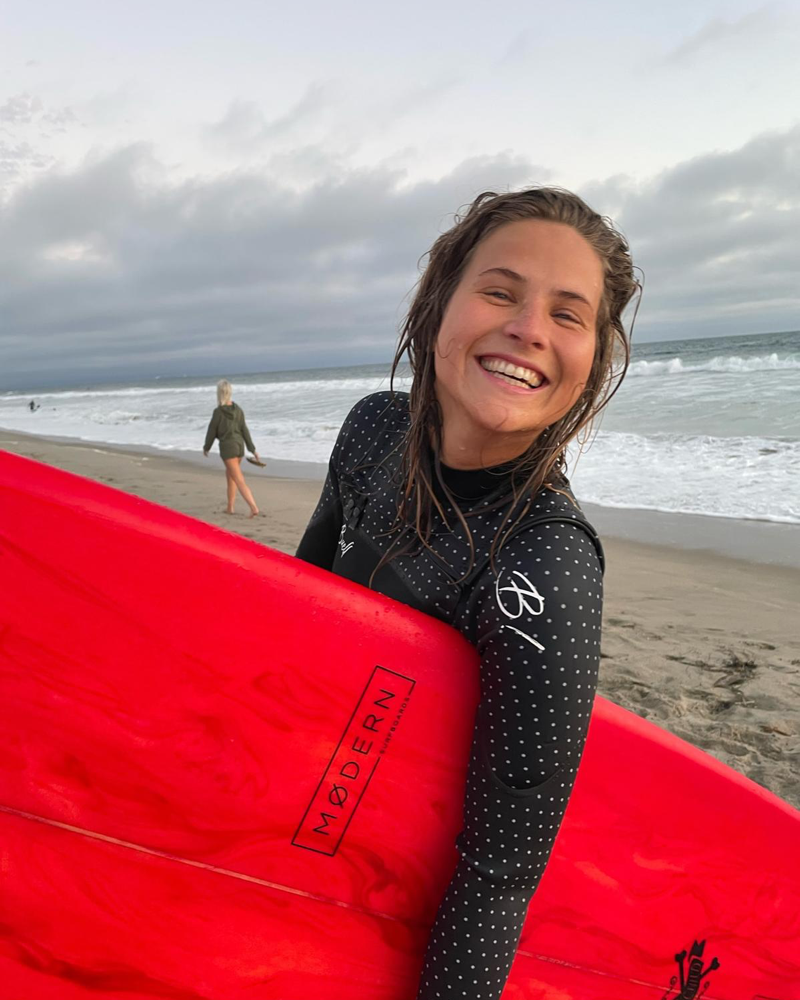 Laura smiling in a wetsuit while holding a bright red surfboard on the beach